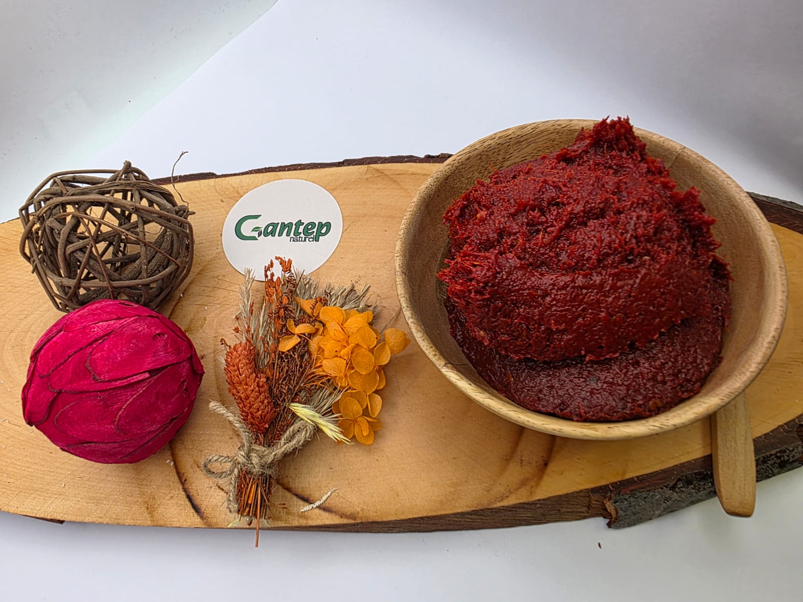Portion scharfer Paprikamark von Gantep Naturel in einer Holzschale, angerichtet auf Holzbrett mit dekorativen Elementen wie Trockenblumen und roter Frucht, aufgenommen auf hellem Hintergrund.