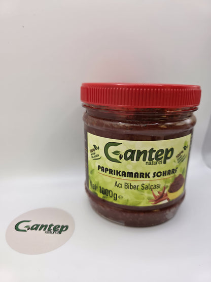 Gantep Naturel scharfer Paprikamark im 1000-g-PET-Behälter mit rotem Schraubdeckel; Frontansicht der Verpackung mit grünem Etikett und Produktdetails, fotografiert auf hellem Hintergrund.