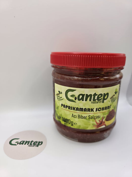 Gantep Naturel scharfer Paprikamark im 1000-g-PET-Behälter mit rotem Schraubdeckel; Frontansicht der Verpackung mit grünem Etikett und Produktdetails, fotografiert auf hellem Hintergrund.