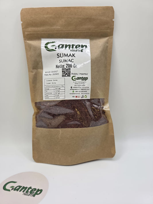 Papiertüte mit Sichtfenster, gefüllt mit 200 g grob gemahlenem Sumak (Sumac) von Gantep Naturel. Die Verpackung zeigt das Markenlogo, Produktangaben auf Deutsch und Türkisch, ein QR-Code sowie Nährwerte. Der Sumak hat eine rotbraune Farbe und ist durch das Fenster gut sichtbar. Im Vordergrund befindet sich ein rundes Etikett mit dem Gantep Naturel Logo.